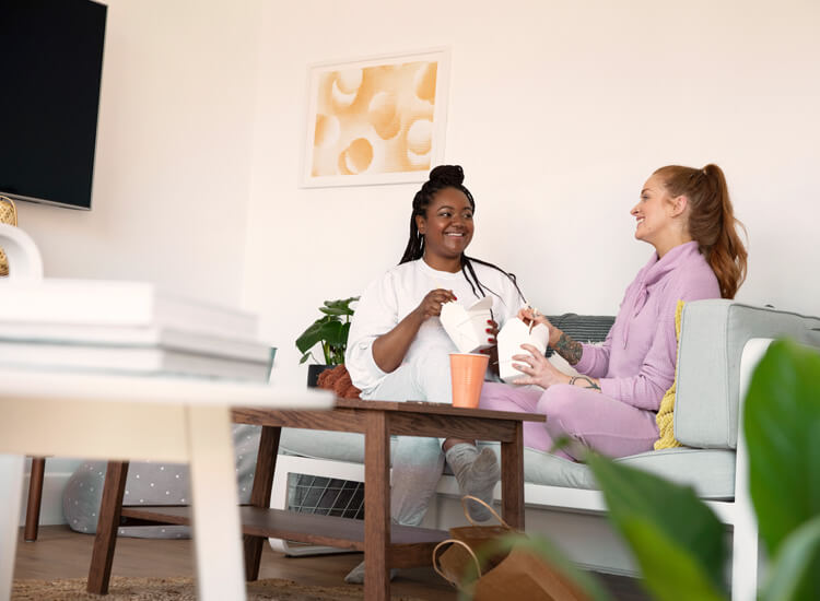 Two women sitting on a sofa at home sharing a meal and talking together