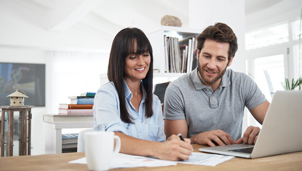 Two people at table, one writing on paper, the other using a laptop, with bookshelves in background.