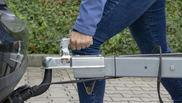 Person attaching trailer hitch to vehicle with greenery in background.